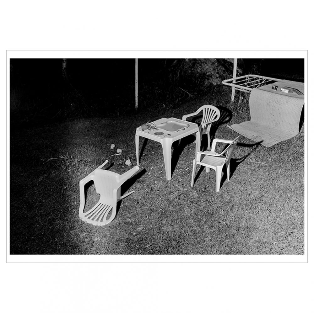 Black and white landscape photograph featuring white plastic garden furniture in the foreground and an outdoor clothes dryer in the back right corner.