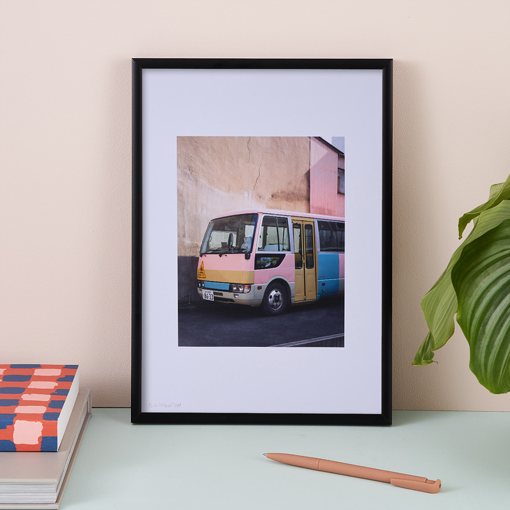 Pink Bus Print by Alice Ishiguro Tosey. The print features a pastel coloured bus against a peach coloured wall. The print is framed in a black wooden frame. 
