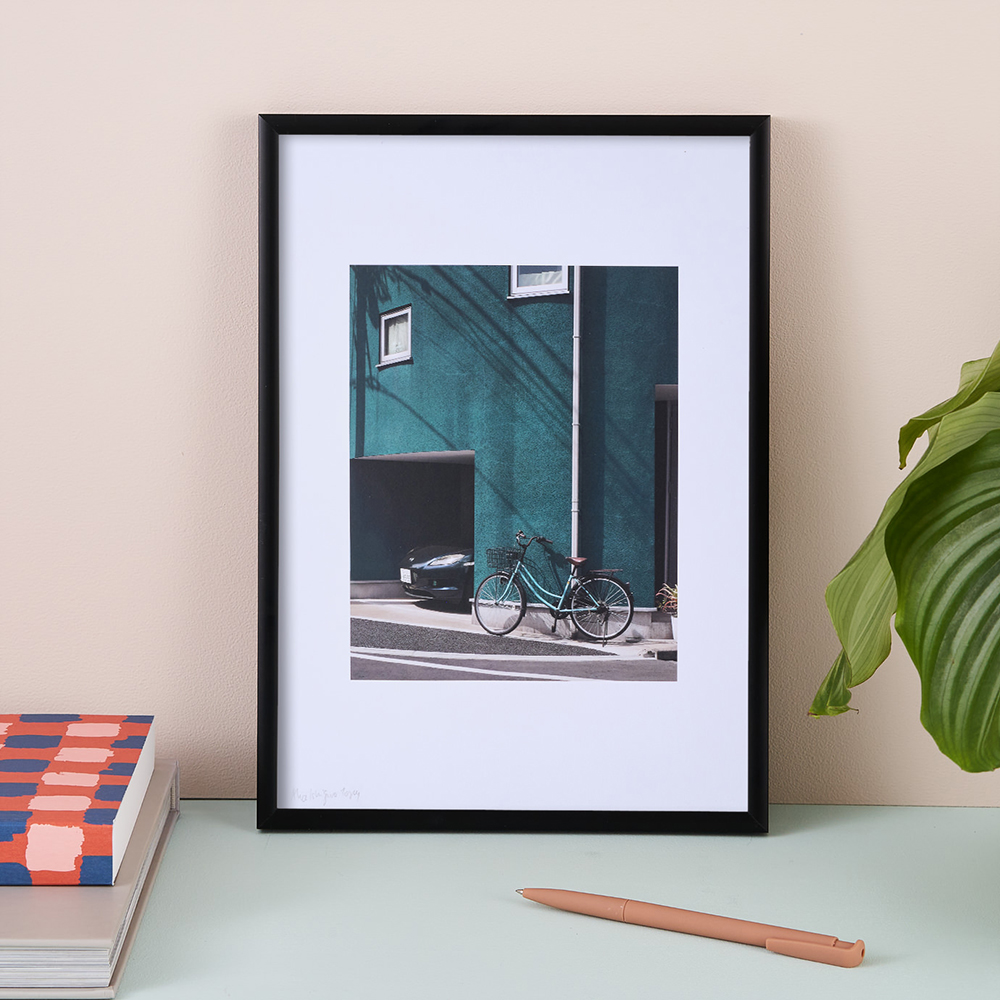 Green building Print A3 by Alice Tosey. The photographic print features a bicycle and black car next to a teal wall with a large white border. The print is framed in a black, wooden frame.