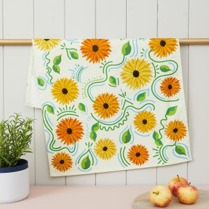 A cotton tea towel, with a cream background colour. Decorated with illustrations of yellow and orange sunflowers and green leaves and foliage in between. The tea towel is draped over a golden rail, with a rosemary herb put to its left, and a pile of three apples to its right.