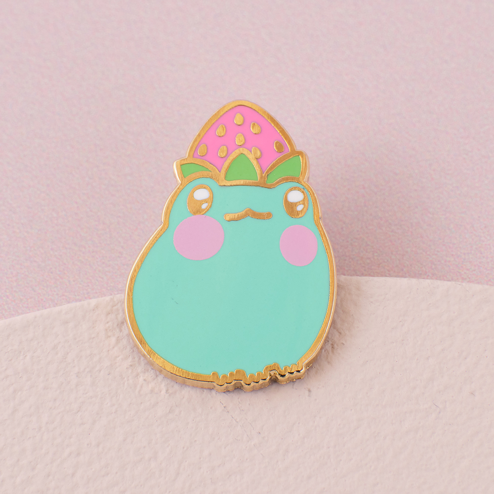 Enamel pin of a green frog with a pink strawberry balanced on it's head