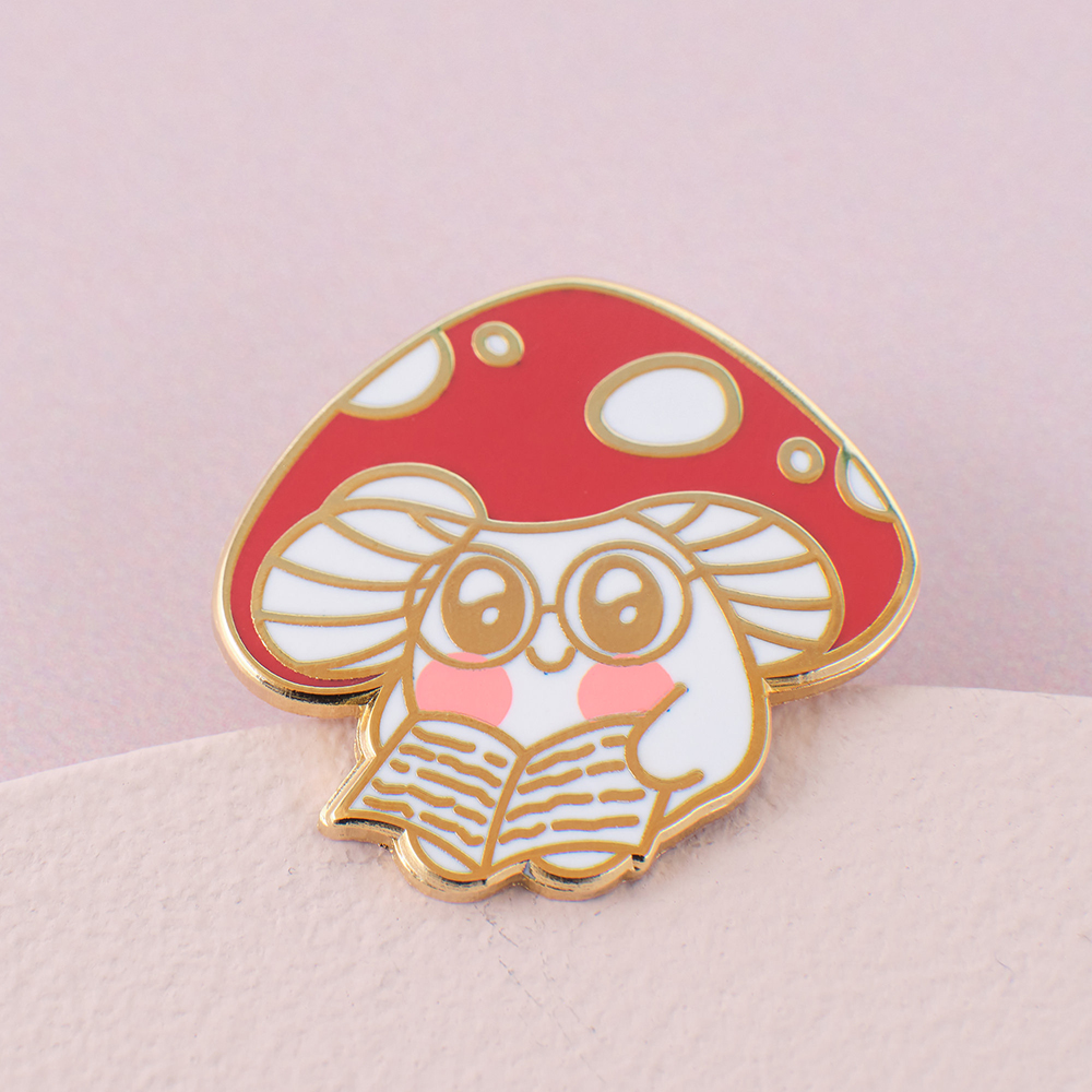 enamel pin badge of a mushroom reading a book on a pink card background