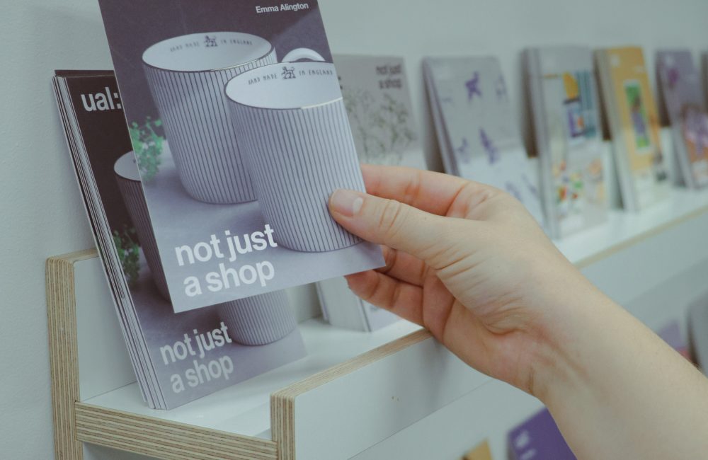 "A hand reaches out to pick up a flyer from a neatly arranged display shelf. The flyer, branded with 'UAL: not just a shop,' features an image of striped ceramic mugs and the name 'Emma Alington.' Surrounding the flyer are other colorful designs and promotional materials for various products, creating an organized and inviting display."