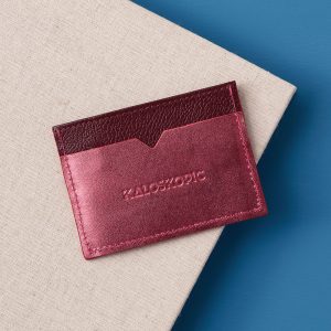 The back panel of a leather card holder in metallic cherry and burgundy reds. The brand name, Kaloskopic, is embossed in the middle of the back panel.