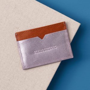 The back side of a leather card holder in metallic lilac and grainy tan. The brands logo, Kaloskopic, is embossed int eh middle of the back panel.