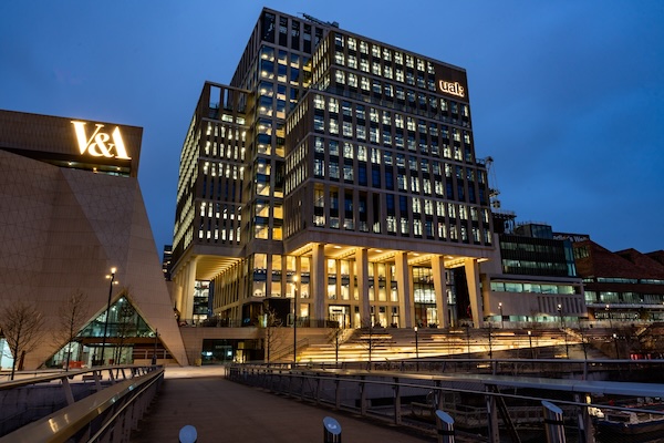 A night view of the London College of Fashion building at East Bank, Stratford, with the V&A Museum building visible on the left. The modern architecture features illuminated windows and tall columns, giving a warm glow against the evening sky. The UAL (University of the Arts London) logo is lit up on the upper part of the building. The pathway leading to the entrance is lined with trees and lights, creating a welcoming atmosphere.