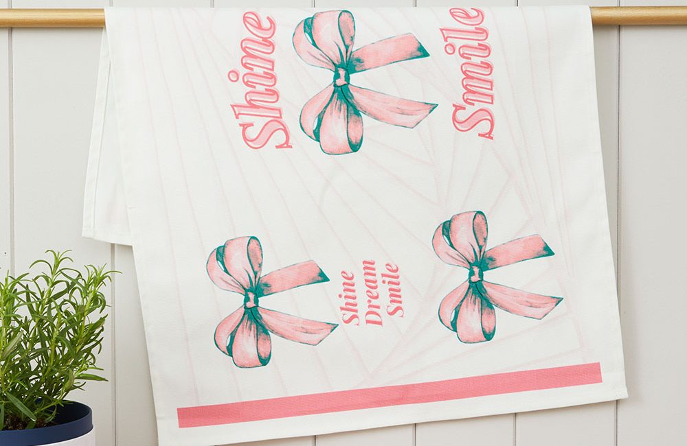 A white tea towel, featuring five pink bows with long tails. Amongst the bows the words, Smile, Shine, Dream are written in pink.