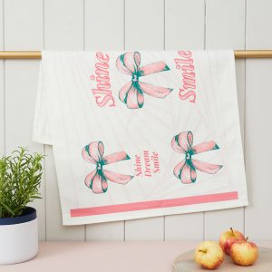 A white tea towel, featuring five pink bows with long tails. Amongst the bows the words, Smile, Shine, Dream are written in pink.