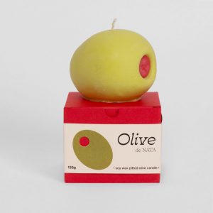 A soy wax candle in the shape of an oversized green olive on top of a red presentation box with a cream belly band with illustrations of the candle and logo.