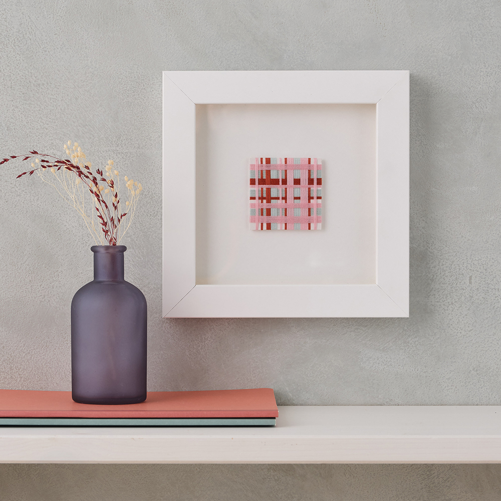 Woven pink, red and blue small square artwork. Mounted on grey wall.