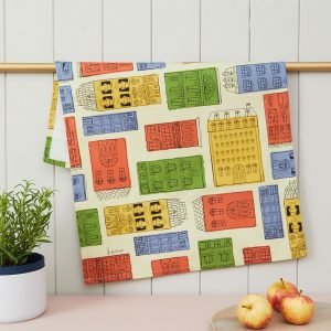 A cotton tea towel with drawings of house fronts inspired by Notting Hill, filled in with primary colours. The tea towel is draped over a golden rail, with a rosemary herb put to its left, and a pile of three apples to its right.