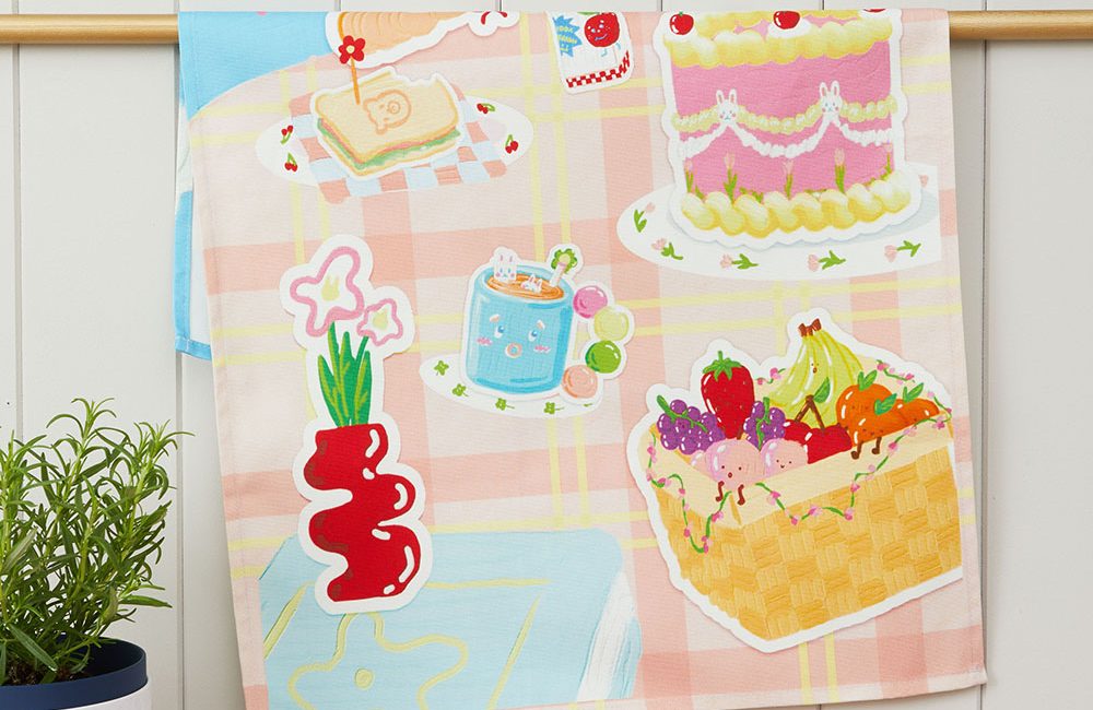 A colourful and childlike illustration on a tea towel. In the style 3d effect style stickers, featuring a cake and fruit basket. Draped over a gold rod in a kitchen setting.