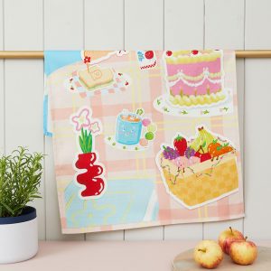 A colourful and childlike illustration on a tea towel. In the style 3d effect style stickers, featuring a cake and fruit basket. Draped over a gold rod in a kitchen setting.