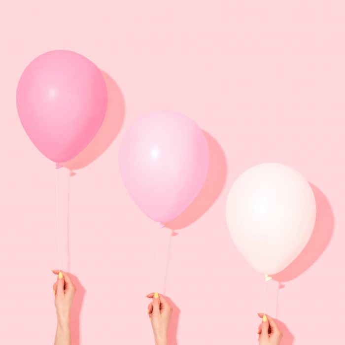 Two pink balloons and one white balloon on a pink background held up by hands