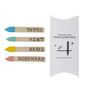 Four slim rectangular ceramic labels with pointed ends. Each one has a different herb name on them: thyme, mint, basil and rosemary. One end of each stick is pointed and painted in a different colour: blue, green, yellow and pink. Next to the sticks is a pillow box packaging in white,, made out of basil seed paper.